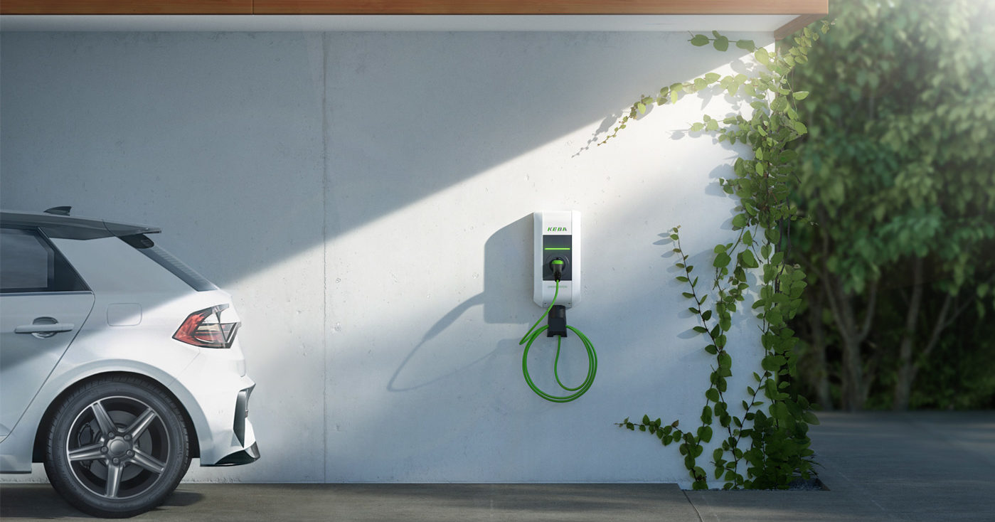 Keba P30 sähköauton latausasema vaalealla sienällä, latauksessa vaalea auto, vihreä iso pusikko latausaseman vieressä.