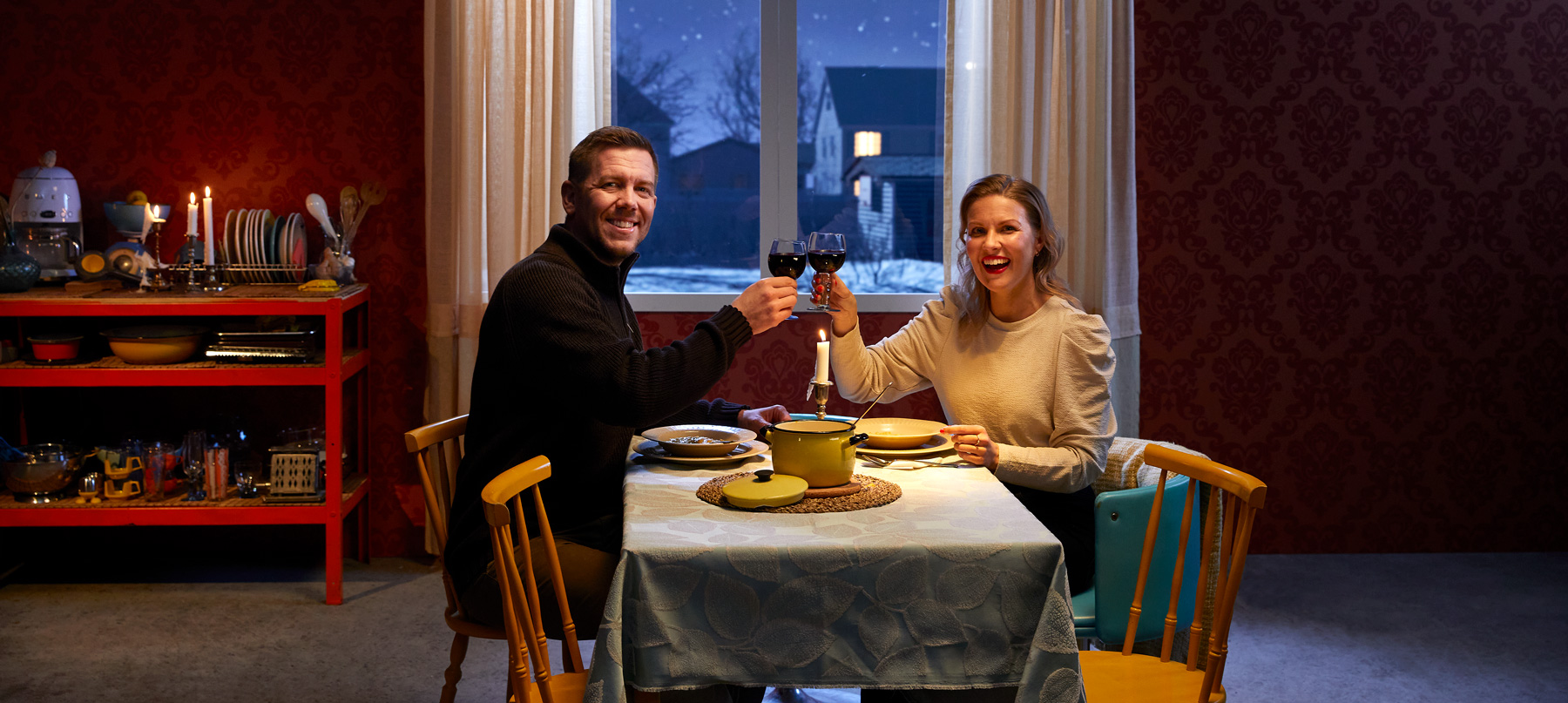 Mies ja nainen ruokapöydän äärellä viettämässä vuosipäivää. Molemmat kohottavat maljaa ja katsovat kameraan.