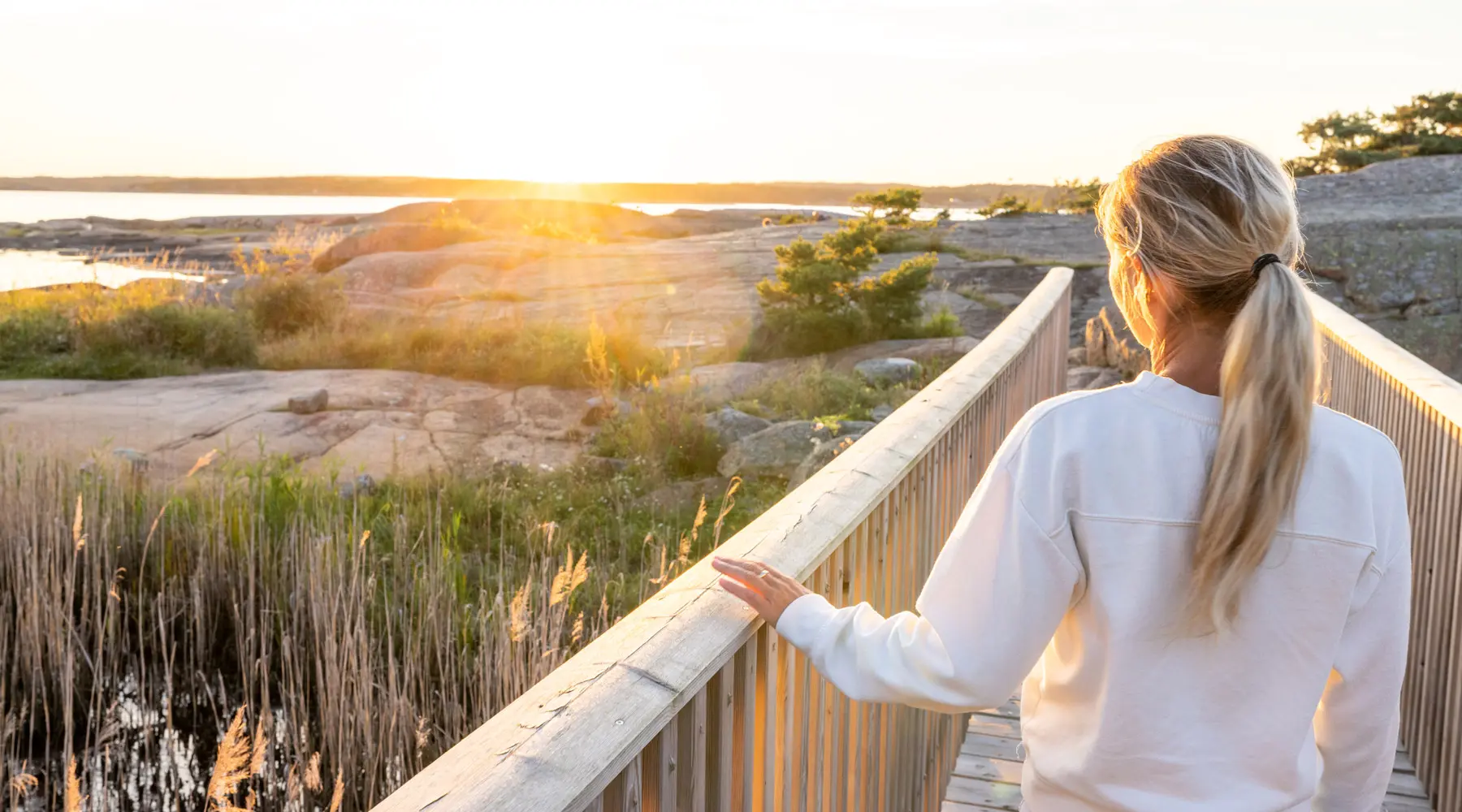 Nainen katsoo kallioiselta paikalta merelle. Aurinko hieman häikäisee katsojaa.