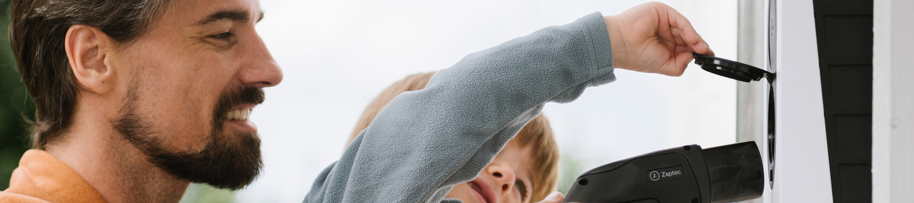 Lapsi kytkee isän avustaessa sähköauton latausasemaan, joka on varustettu dynaamisella kuormanhallinnalla, tehostaen latauksen hallintaa.