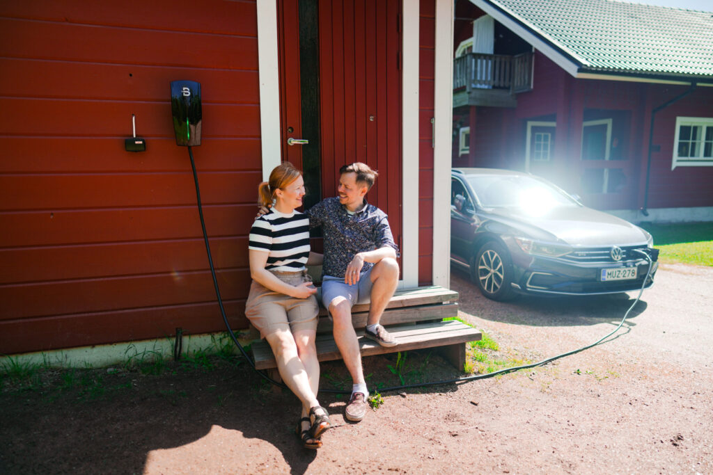 Pariskunta istuu portailla samalla, kun sähköauto latautuu Beny sähköauton latausasemalla